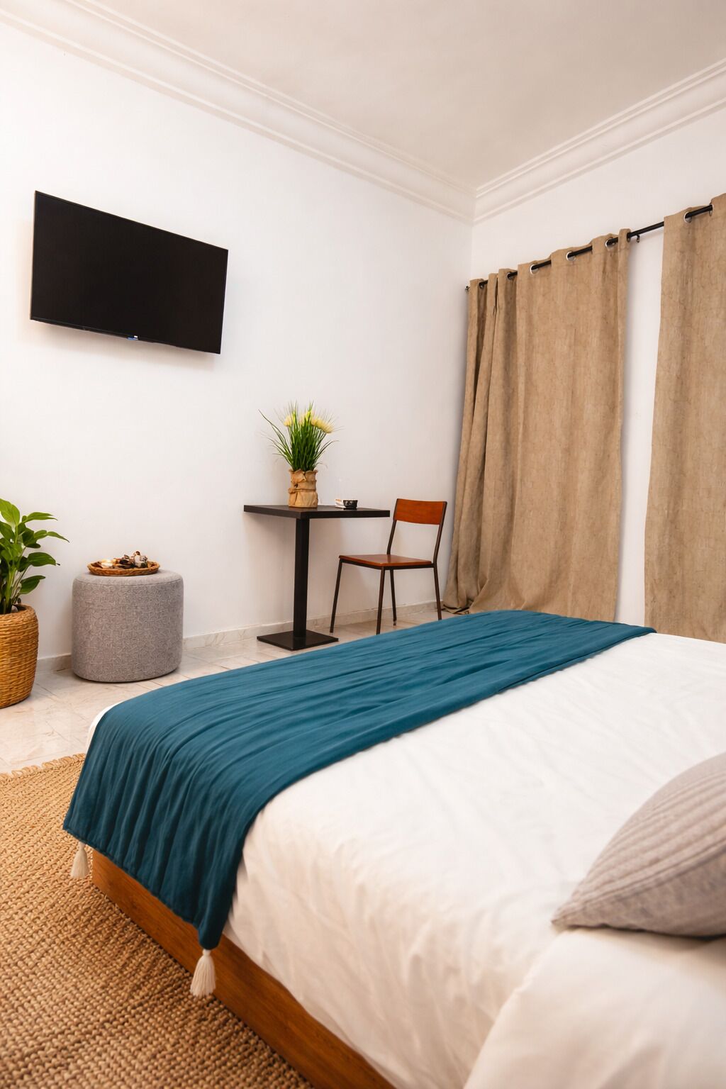 A bedroom with a bed, a TV mounted on the wall, a desk with a chair, a potted plant, and a window with curtains.