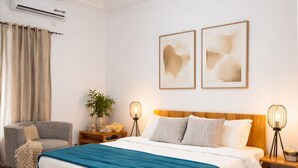 Um quarto com cama de madeira, uma poltrona cinza, uma mesa de cabeceira com abajur e ar condicionado fixado na parede.