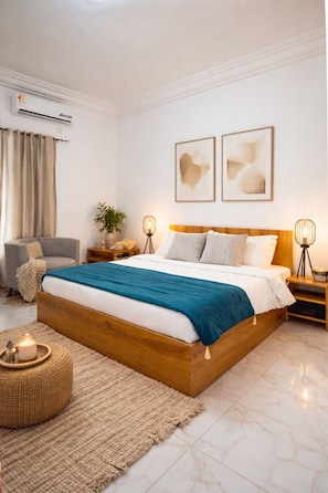 A bedroom with a wooden bed, a grey armchair, a bedside table with a lamp, and a wall-mounted air conditioner.