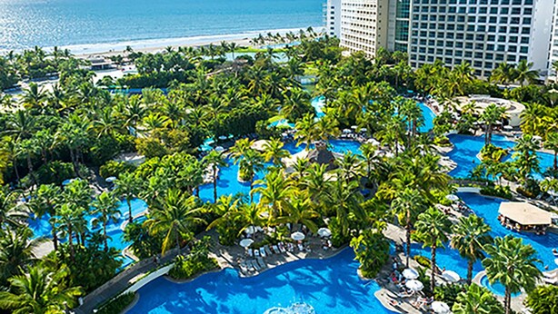 A resort with multiple swimming pools surrounded by palm trees, a beach, and a large hotel building.