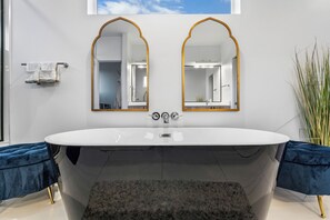 A modern bathroom with a freestanding bathtub, two large mirrors with gold frames, a towel rack, and a potted plant.