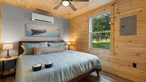 Ein schlicht eingerichtetes Schlafzimmer mit Holzwänden, einem Bett, zwei Nachttischlampen, einer Klimaanlage, einem Deckenventilator und einem Fenster mit Blick auf eine grüne Landschaft.
