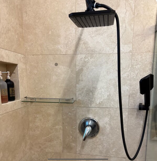 Une douche équipée d’un pommeau de douche à main et d’une commande fixée au mur. On y trouve une étagère en verre pour les articles de toilette et un porte-serviettes.