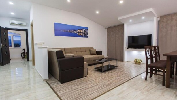 A modern living room with a sofa, coffee table, and TV. There is a dining area with chairs and a table. The room has a large window and an air conditioning unit on the wall.