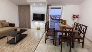A modern living room with a sofa, a coffee table, a TV mounted on the wall, a dining table with chairs, and a balcony with a view.