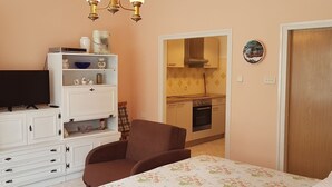 Une chambre avec un lit, un téléviseur, un fauteuil marron et, au fond, une petite cuisine.