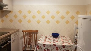 Une cuisine avec une chaise en bois, une table recouverte d’une nappe à motifs floraux, un bol bleu et un réfrigérateur.