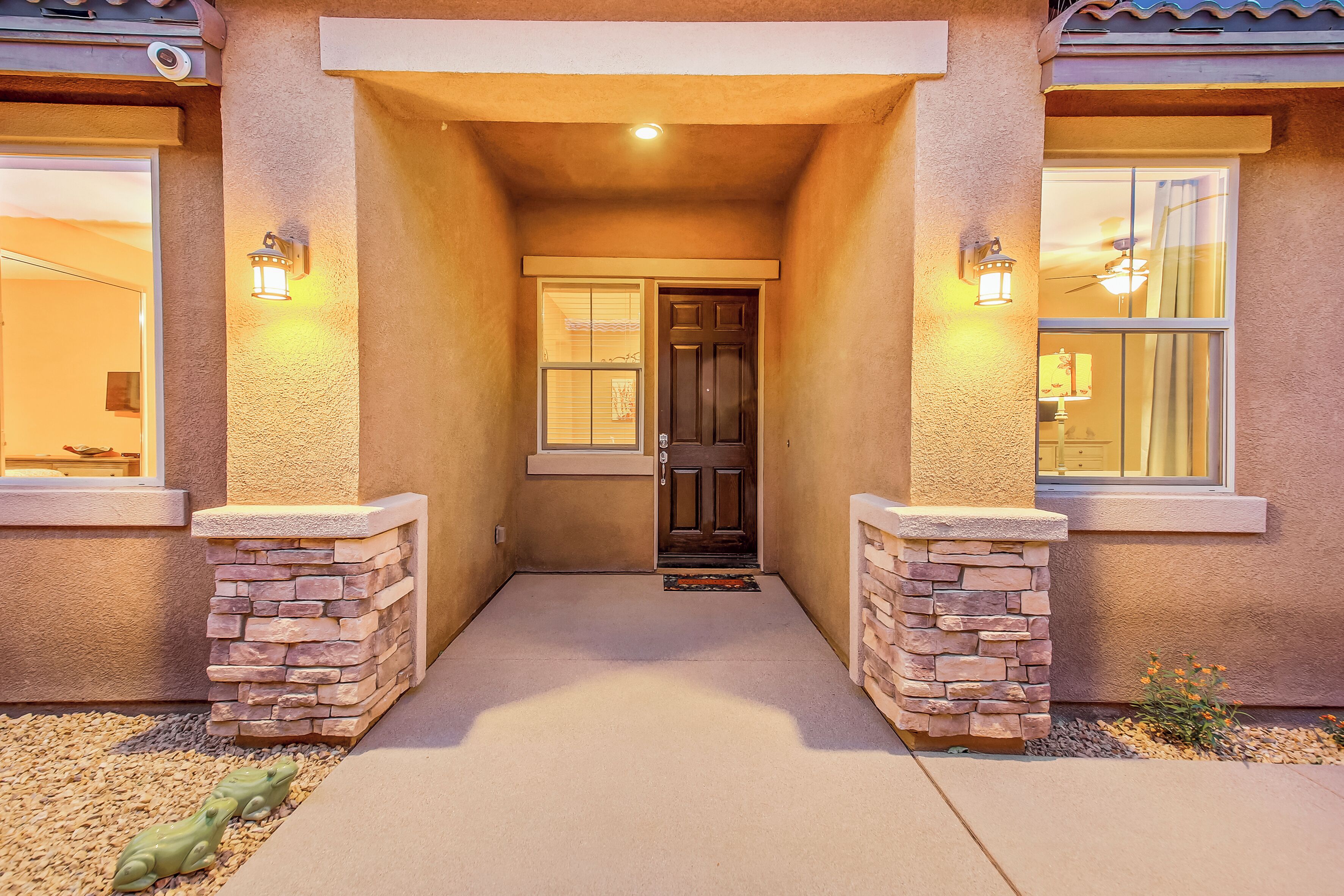 A house entrance with a concrete pathway, a dark door, stone pillars, and two lit wall sconces.