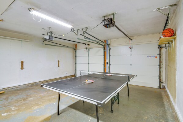 Een tafeltennistafel in een ruimte met een garage deur, een basketbal, en zichtbare water- en elektriciteitsbuizen.