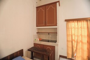 A compact room with a bed, a small table, a kitchenette with a sink and cabinets, and a window with curtains. - Mango Tree Retreat (Galewela)