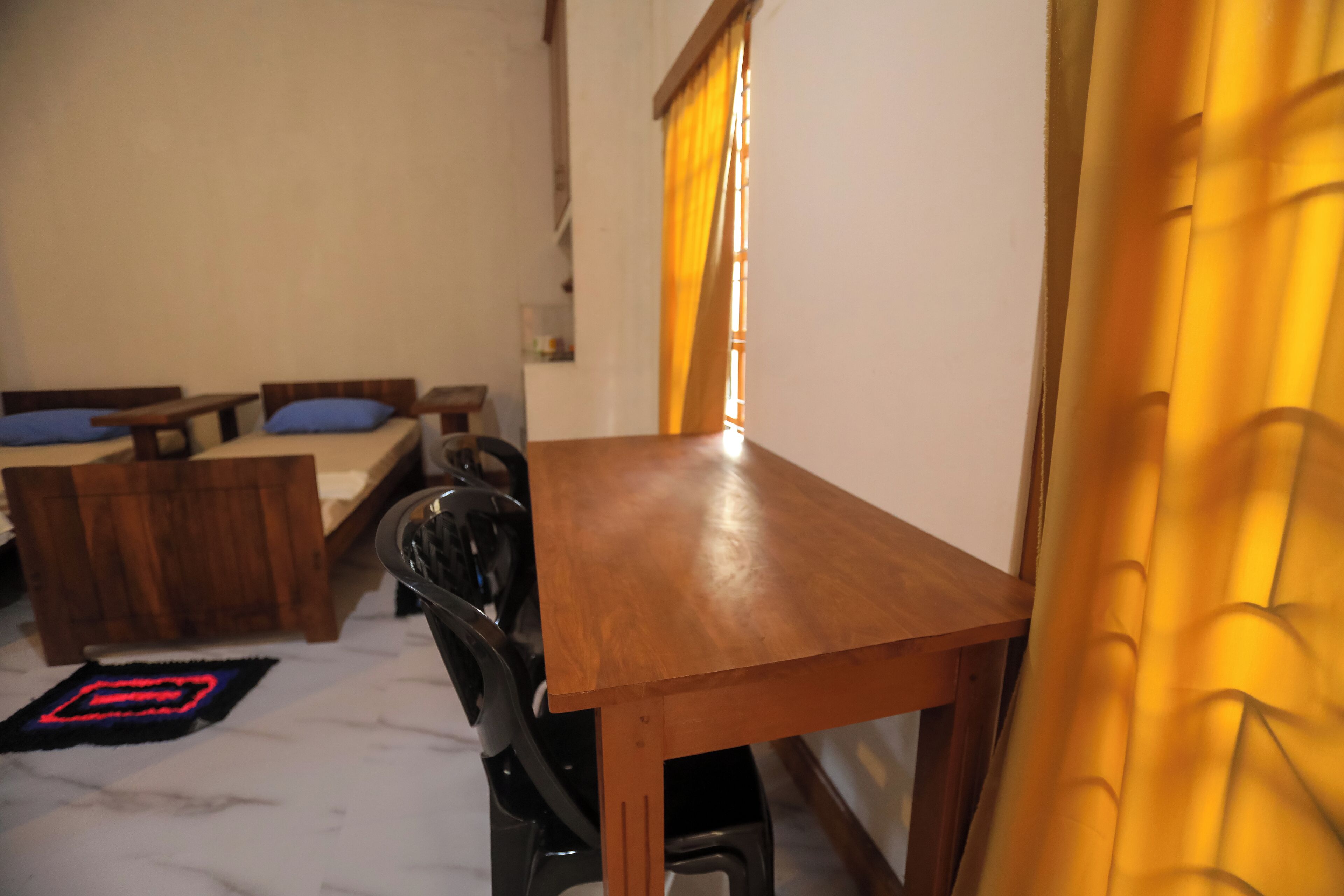 A room with a wooden table, two beds with blue pillows, a black chair, and a yellow curtain.