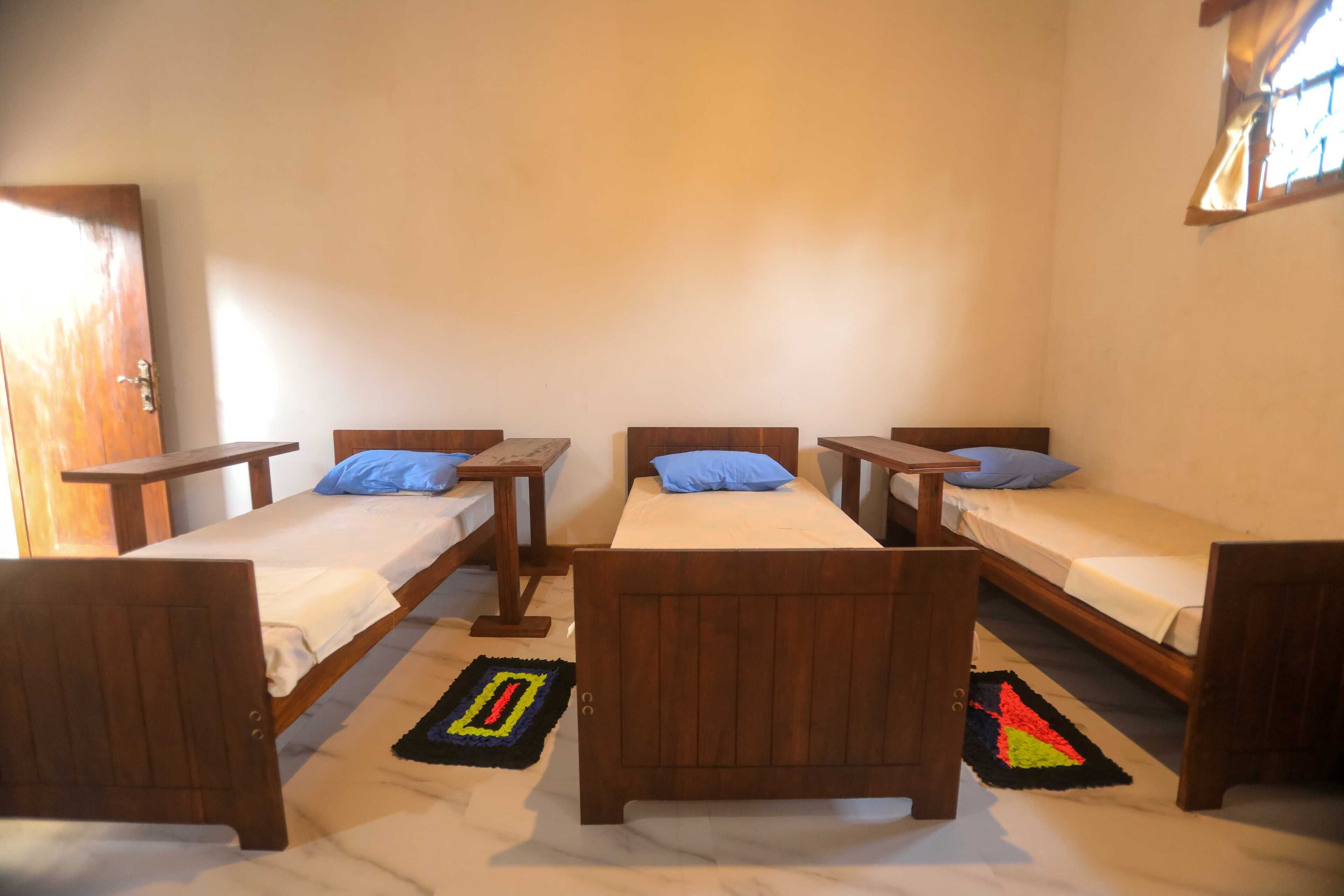 Une pièce comprenant quatre lits en bois disposés en rangée, chacun ayant une tête de lit et un pied de lit en bois assortis. De petites tables de chevet en bois, surmontées de lampes, se trouvent à côté de chaque lit, et un oreiller bleu est posé sur chacun d’eux. La pièce présente un aménagement simple et épuré, avec des murs blancs et un plancher en bois.
