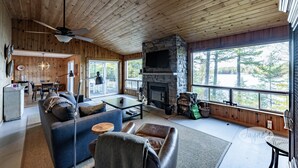 A cozy cabin interior with a stone fireplace, wooden ceiling, and large windows offering a view of the forest. - Darling 3 bed 2 bath cottage on Lake Joseph with Bunkie! NW Exposure! Boathouse (Seguin)