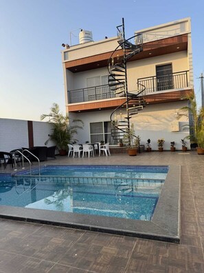A swimming pool with a modern outdoor seating area, a two-story building with a spiral staircase, and a water tank on the roof. - Paradise Villa (Devanahalli)