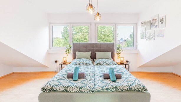 A bedroom with a large bed, two bedside tables, a window with a view of greenery, and a ceiling light fixture.