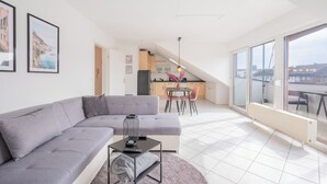 A modern living room with a grey sofa, a small table with a lamp, and a kitchen in the background.