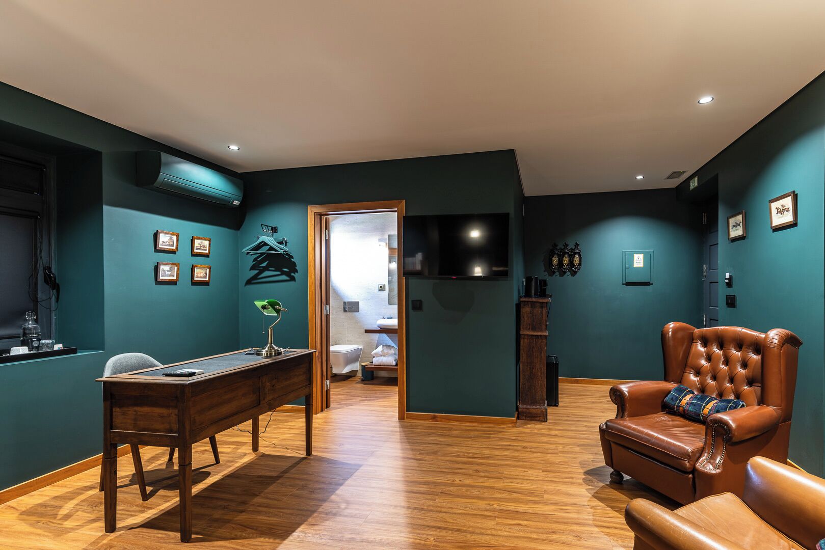 Ein Zimmer mit Schreibtisch aus Holz, einem Ledersessel, teal-farbenen Wänden, einem an der Wand befestigten Fernseher und einem Holzboden.