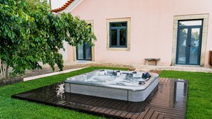 Ein Whirlpool in einem Garten mit einer Holzterrasse, einem Kiesweg und einem Haus mit rosafarbenen Wänden und blauen Türen.