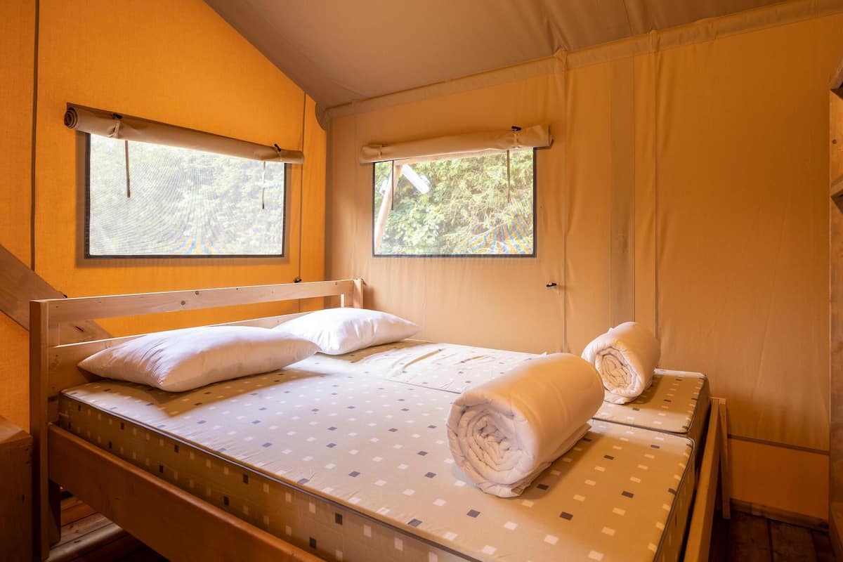 A single bed with a wooden headboard, two windows with curtains, and a small table with a rolled towel.