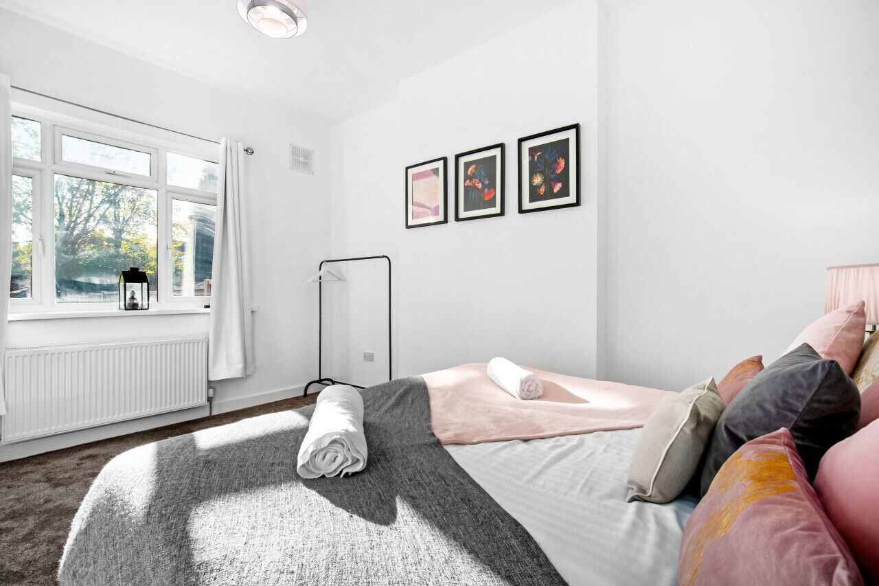 Une chambre avec une grande fenêtre, un radiateur, un lit sur lequel est posée une serviette enroulée, et trois tableaux encadrés accrochés au mur.