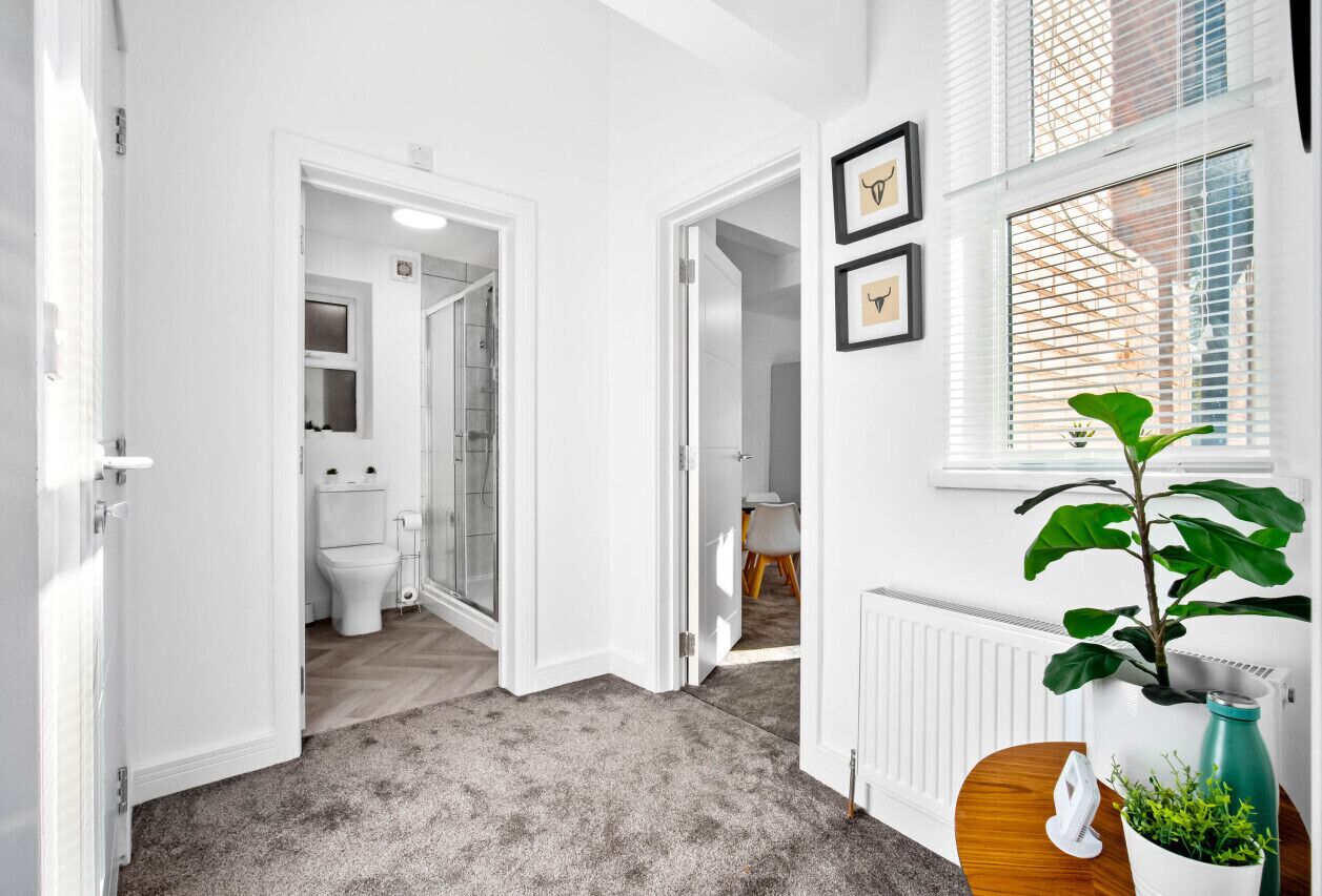 A hallway with a grey carpet, white walls, and a door leading to a bathroom. There is a radiator, a potted plant, and a framed picture on the wall.