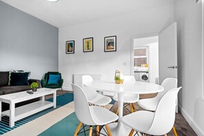 Ein modernes Wohnzimmer mit einem weißen runden Tisch, weißen Stühlen, einem dunkelgrünen Sofa und einem weißen Couchtisch. An der Wand hängen drei gerahmte Bilder.