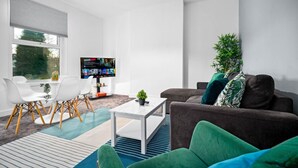Un saló modern amb una taula blanca, un sofà marró, coixins verds i una televisió.