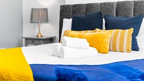 Een net opgemaakt bed met opgerolde witte handdoeken, een blauw-gele deken en decoratieve kussens met verschillende patronen.