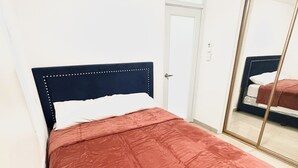 Una cama con cabecera oscura y una manta roja, una puerta blanca y un espejo grande que refleja la cama.