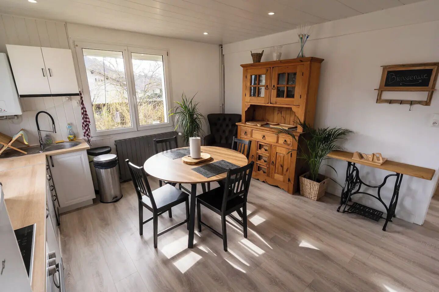 A compact kitchen with a round dining table, wooden cabinets, and a black chair.