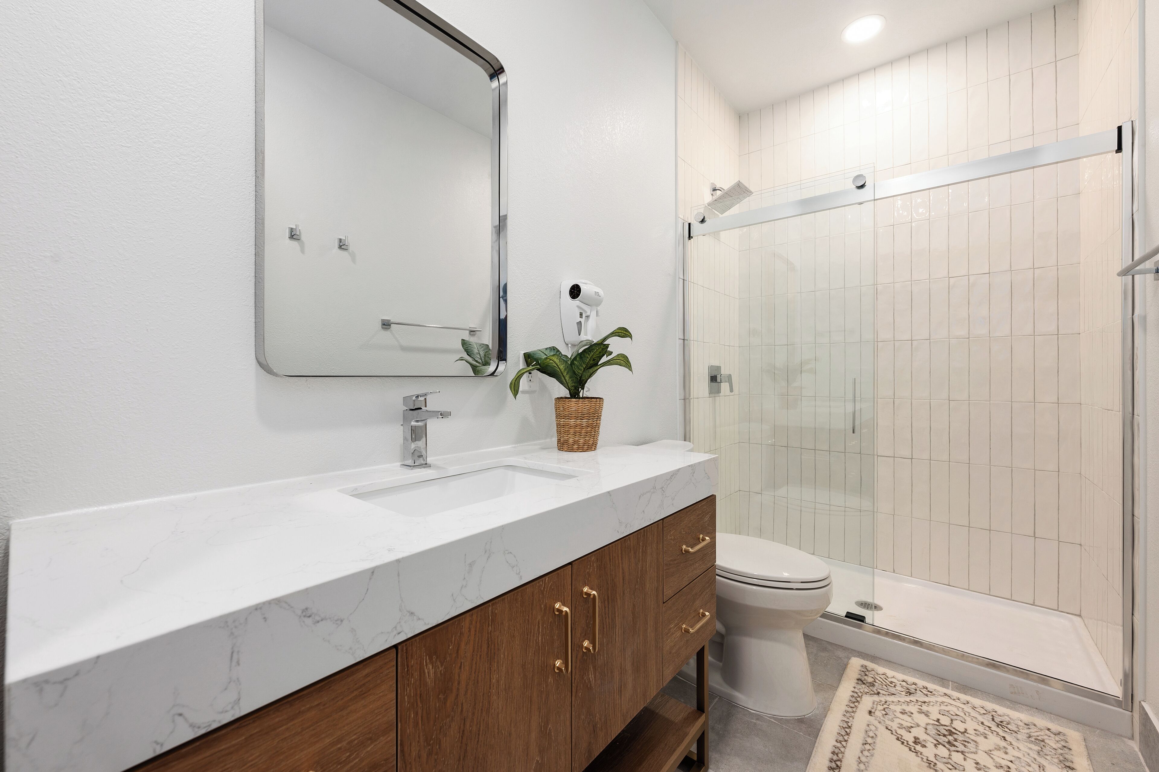 A bathroom with a marble sink, wooden vanity, a mirror, a plant, a shower, and a toilet.