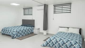 A modern bedroom with two beds, a small bedside table, a grey headboard, and a window with blinds.