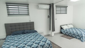 Un dormitorio con dos camas, cabecero gris y ventana con persianas.