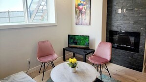 Una sala de estar moderna con televisor, dos sillones rosados, una mesa redonda con un florero, chimenea y un ventanal con vistas a una terraza de madera.