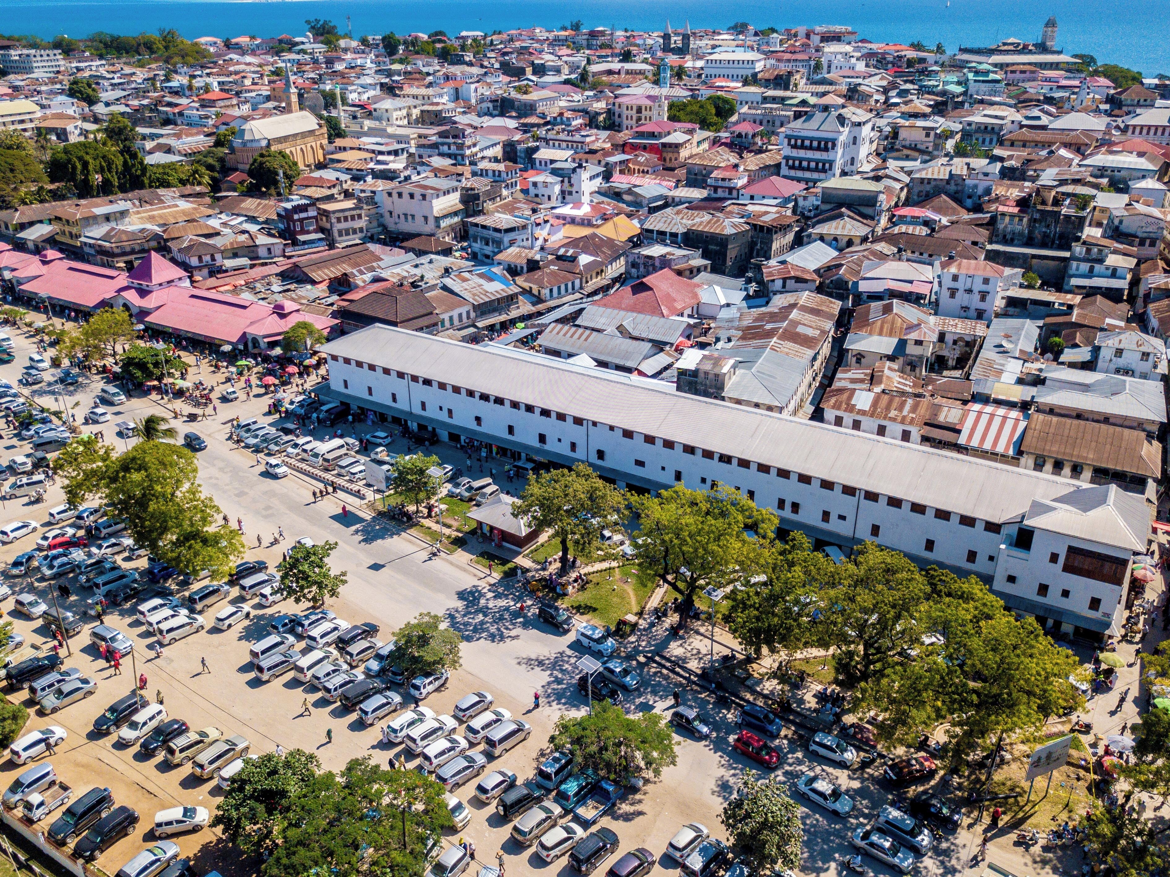 Vista aérea de una zona urbana densamente poblada, con un edificio blanco destacado de techo plano, rodeado de edificios más pequeños y un amplio estacionamiento con diversos vehículos.