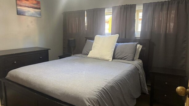 A bedroom with a bed, dark curtains, a nightstand, a lamp, and a painting on the wall.