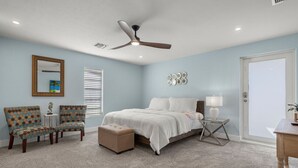 A bedroom with a bed, two chairs, a side table, a ceiling fan, and a door.