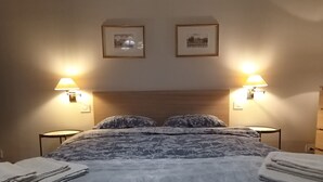 Una cama bien tendida con una colcha estampada y almohadas, dos lámparas fijadas a la pared y dos cuadros enmarcados.