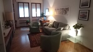 Una sala de estar con un sofá, sillones, una mesa de centro, una lámpara y una planta. Hay una ventana con persianas, una alfombra y un cuadro en la pared.