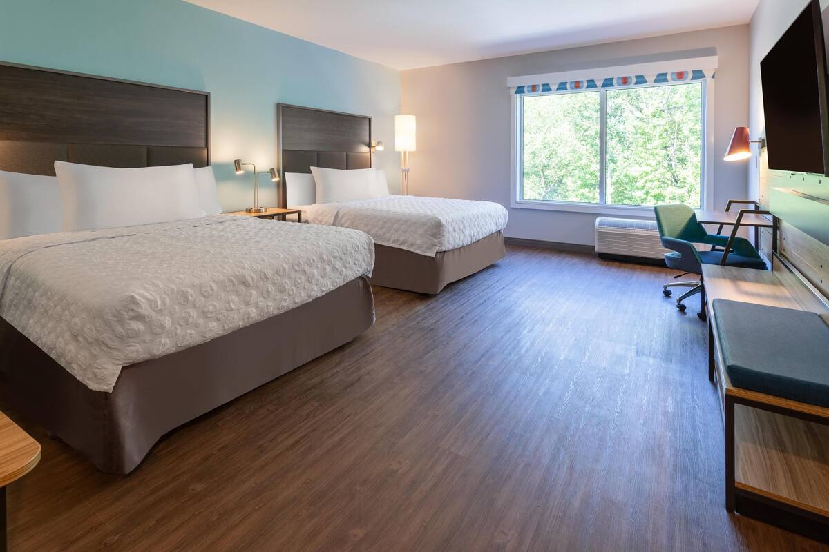 A hotel room with two beds, a desk, a chair, and a large window with a view of trees.