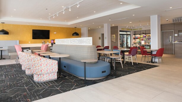 A modern restaurant with a seating area featuring a blue sofa and patterned chairs, a counter with a menu, and a refrigerator stocked with snacks. - Home2 Suites By Hilton Lima (Lima)