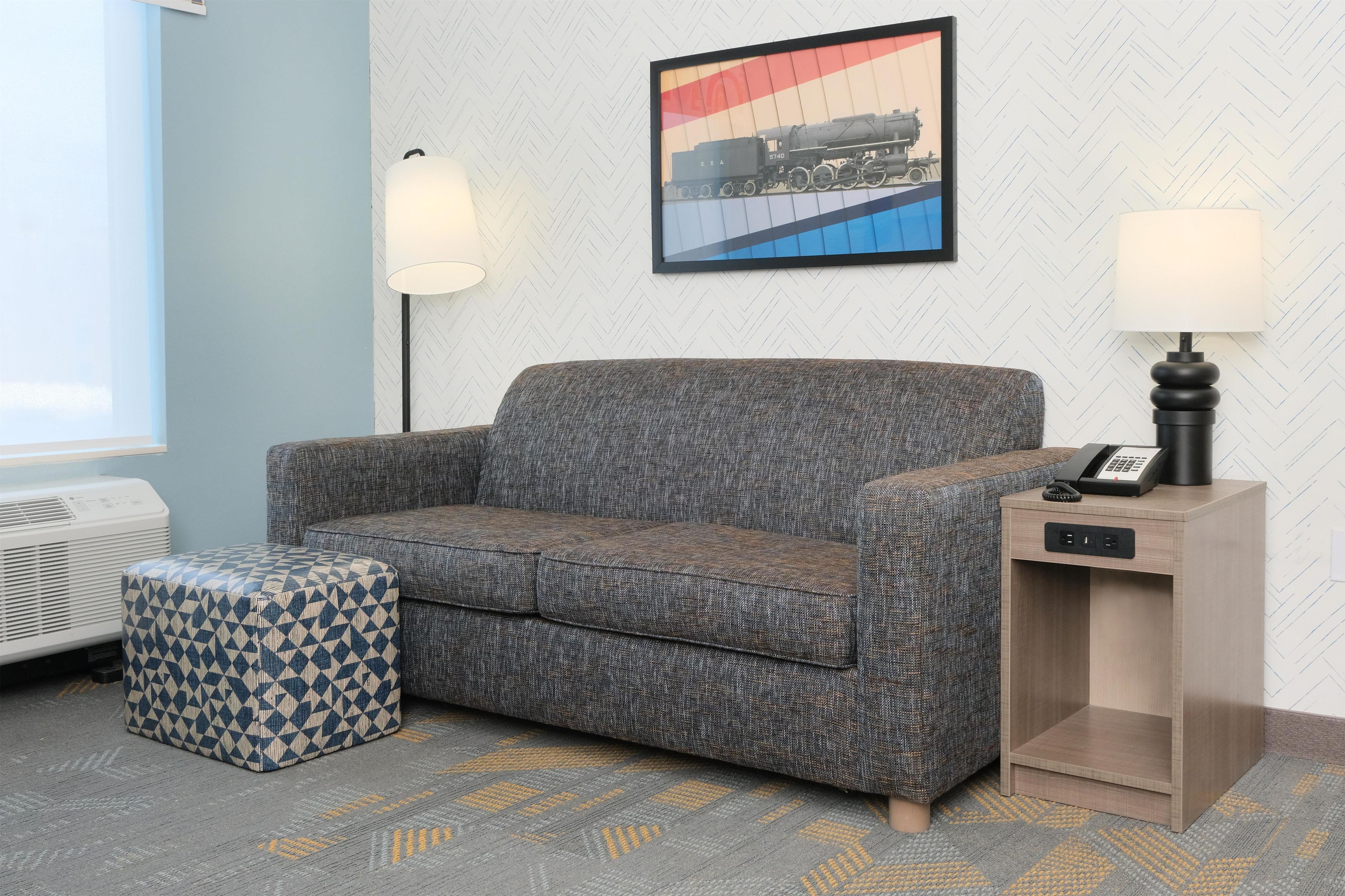 A room with a grey patterned sofa, a matching ottoman, a small wooden side table with a telephone, and a wall-mounted lamp.