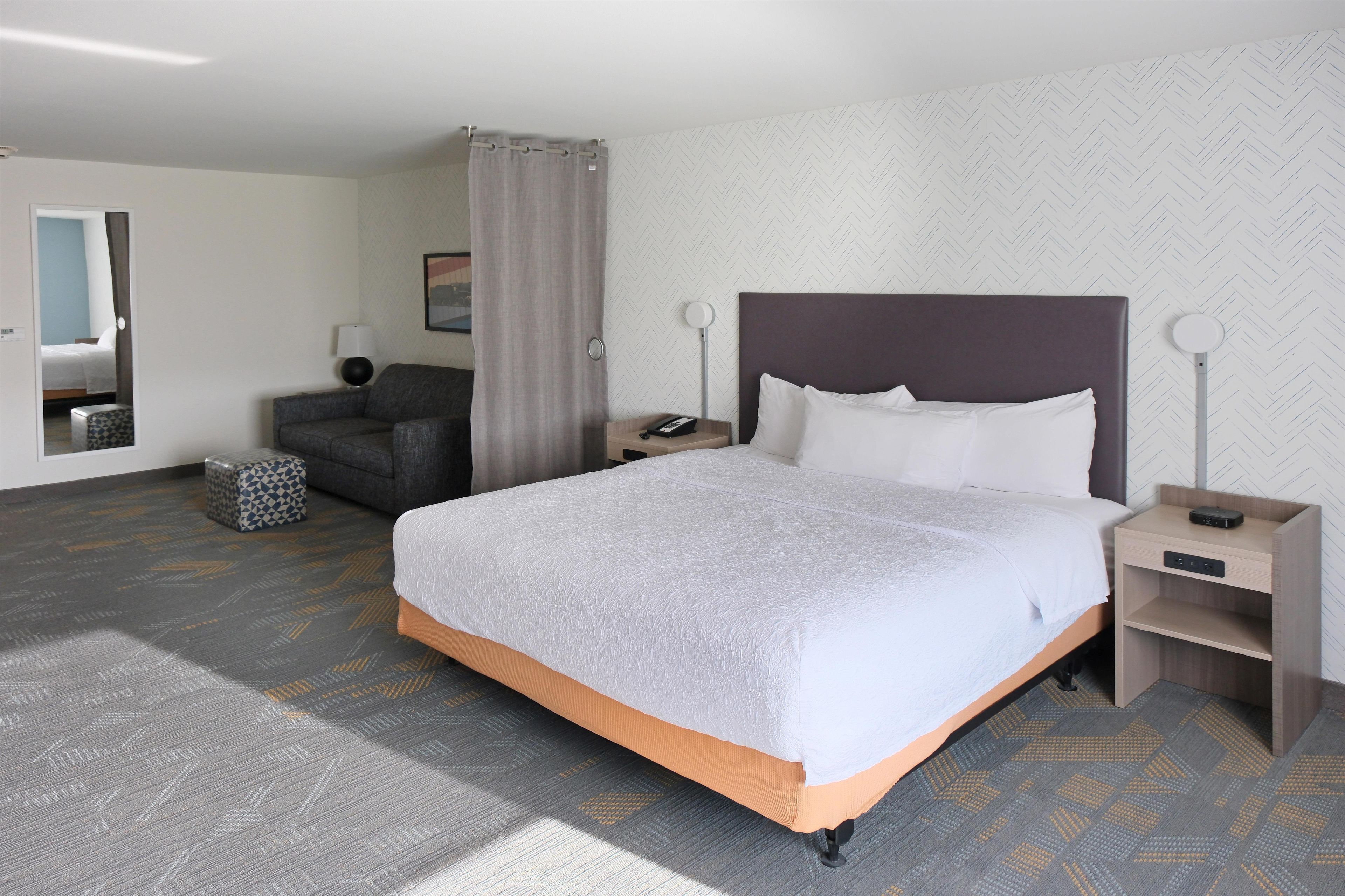 A hotel room with a large bed, a desk, a chair, a nightstand, and a bathroom visible through an open door.