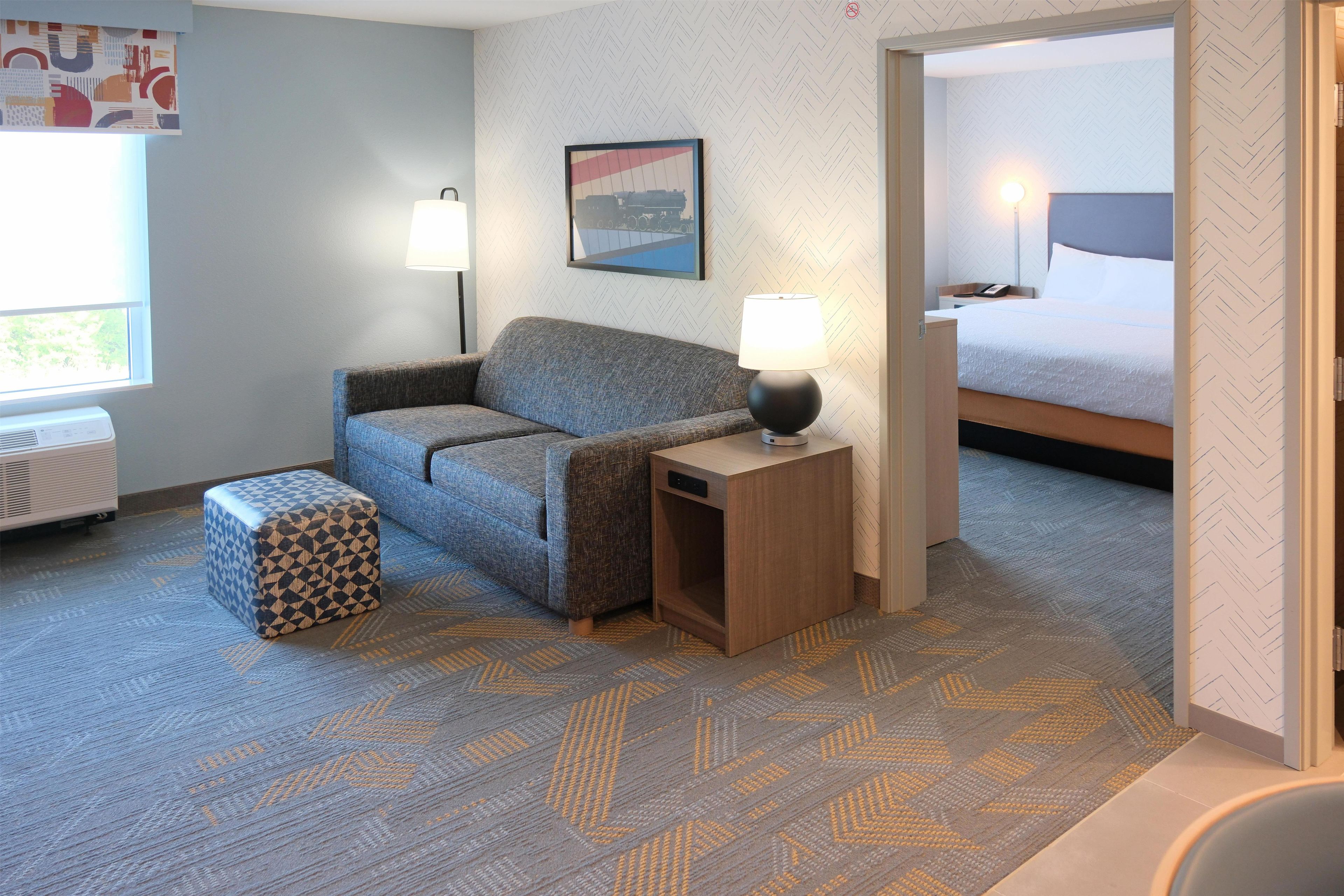 Une chambre d’hôtel avec un canapé, un tapis à motifs, une table de chevet avec une lampe, un lit et une fenêtre donnant sur un paysage.