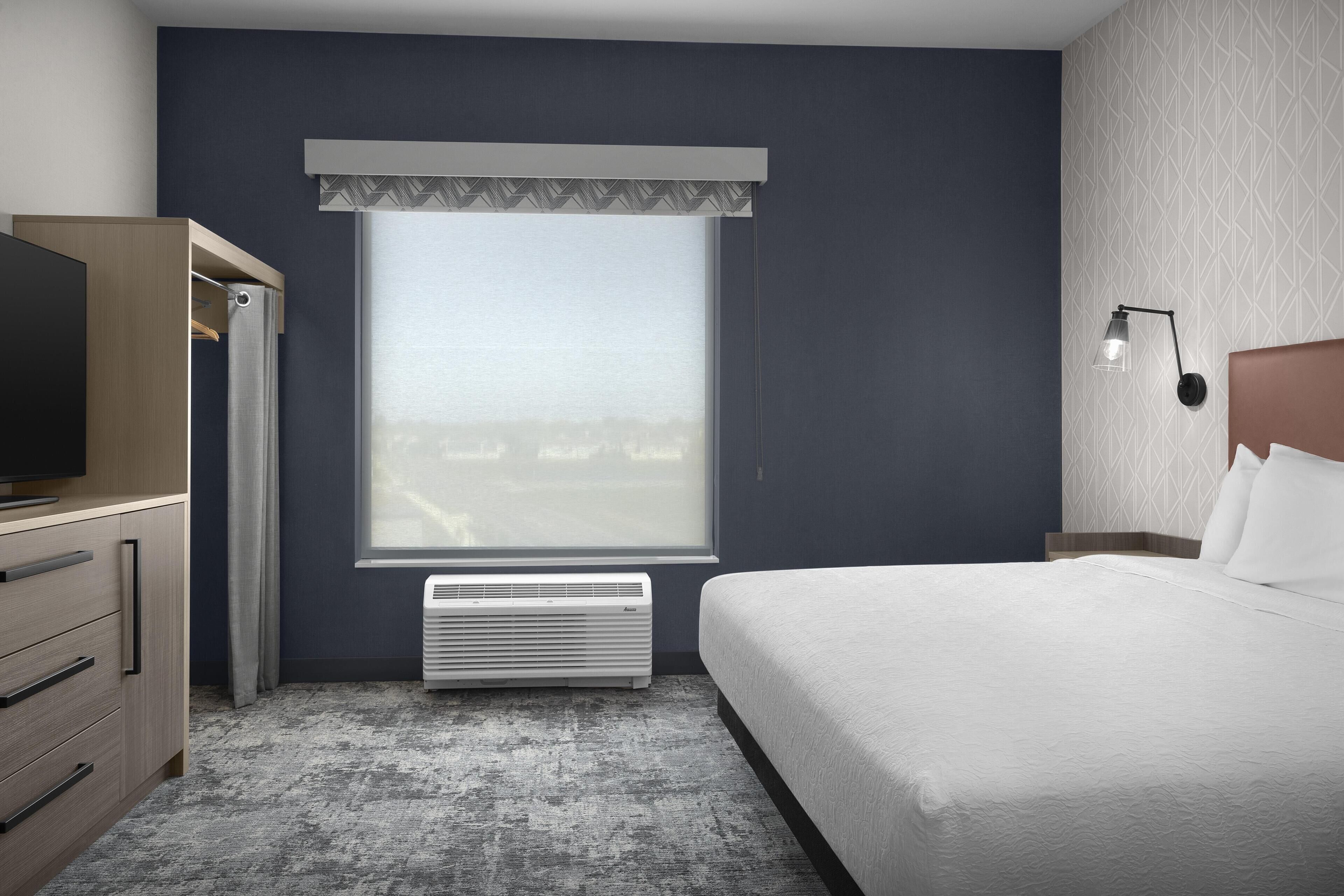 A hotel room with a bed, a TV, a wardrobe, a window with curtains, a wall-mounted lamp, and a radiator.