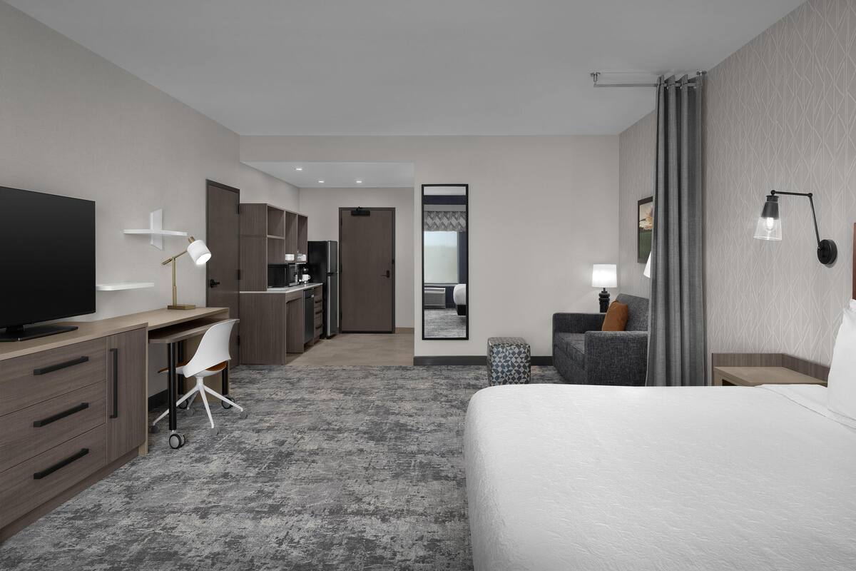 Una habitación de hotel moderna con una cama grande, un escritorio con silla, un televisor y una kitchenette al fondo.
