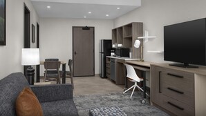Een moderne, goed verlichte woonkamer met een bank, een eethoek met tafel en stoelen, een keuken met een koelkast, magnetron en een televisie.
