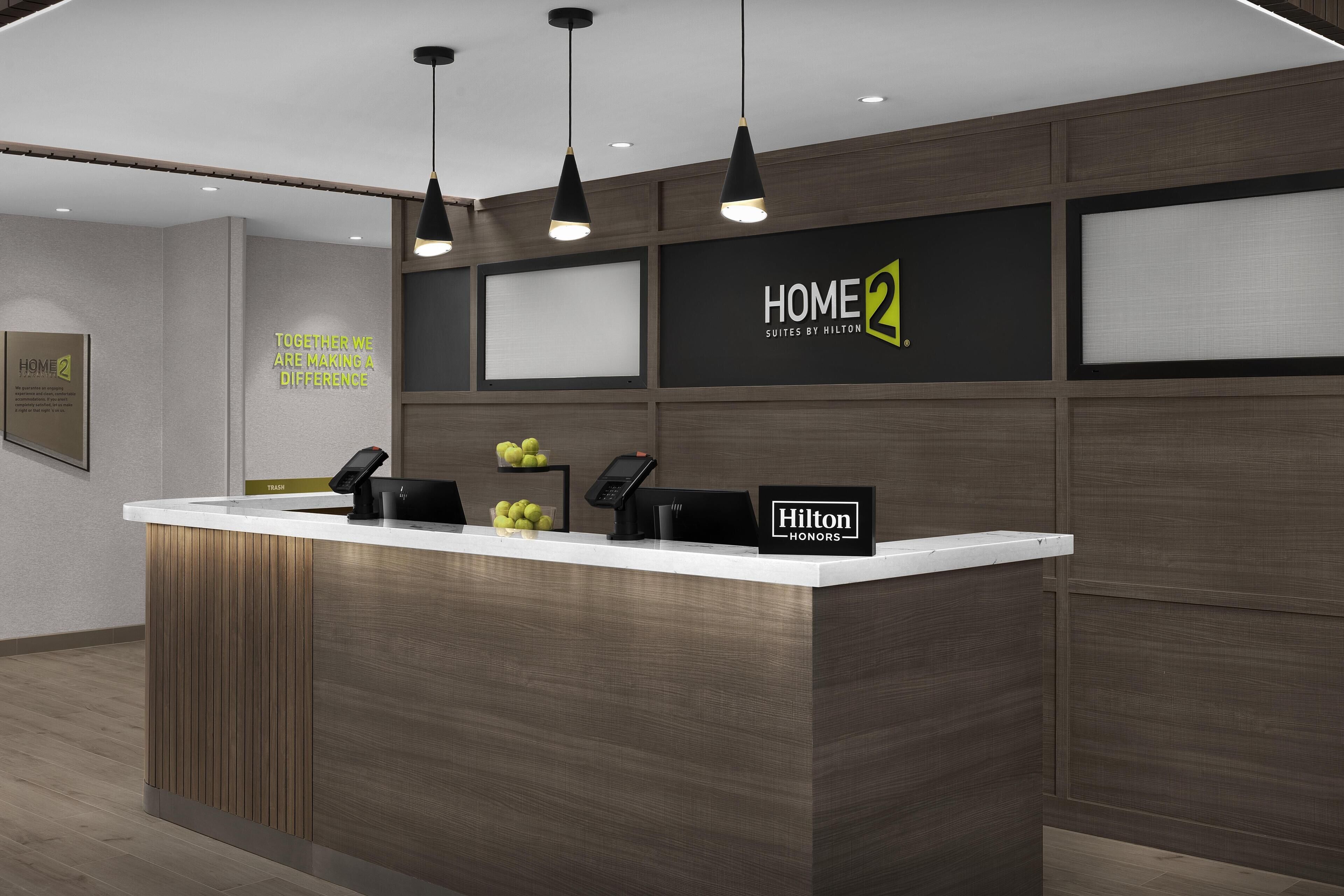 A modern hotel reception desk with a wooden finish, a digital display, and a sign for 'HOME 2 Suites by Hilton'.