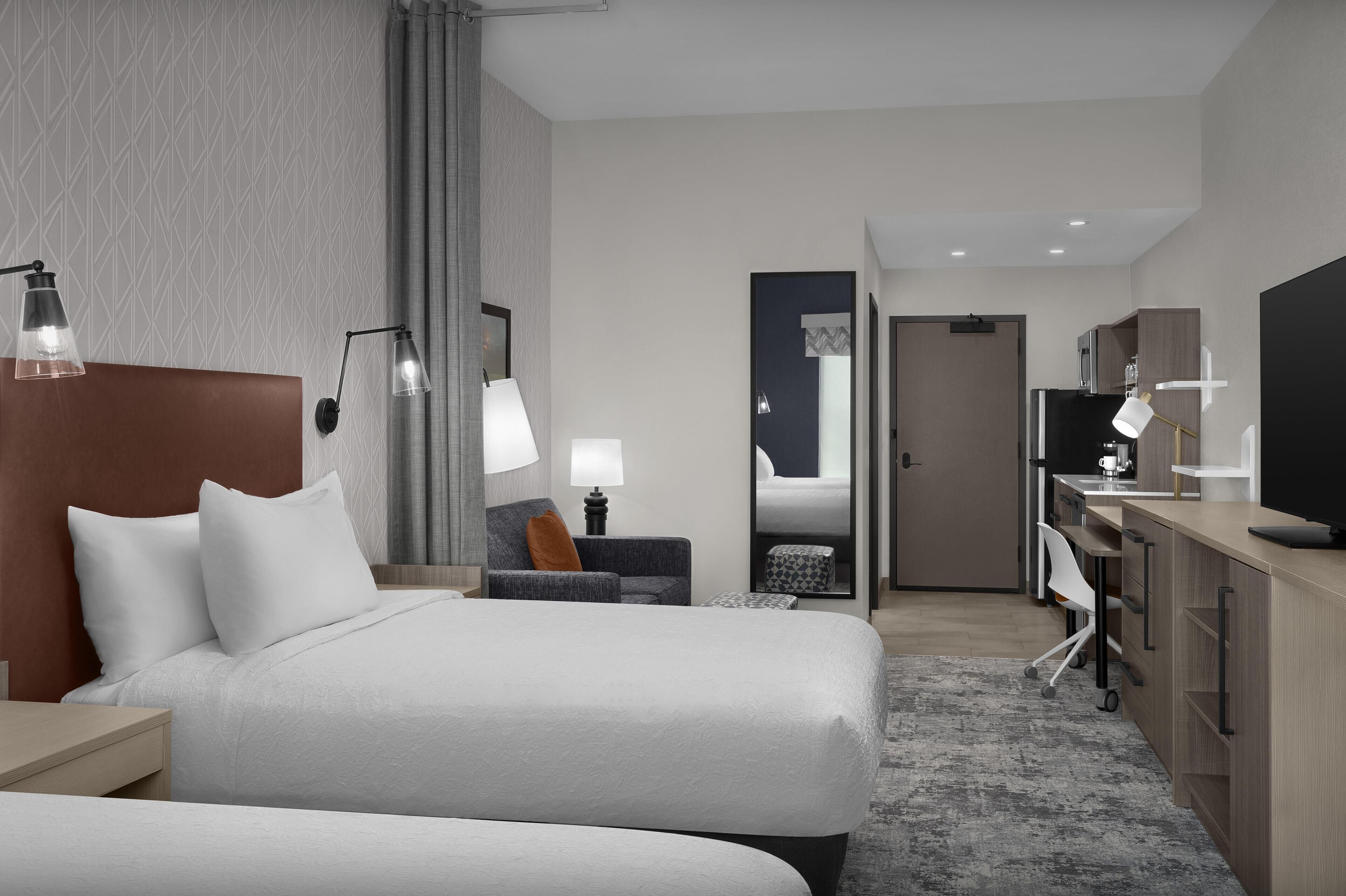 Ein Hotelzimmer mit zwei Betten, einem Schreibtisch, einem Stuhl, einem Fernseher und einem durch eine offene Tür einsehbaren Badezimmer.