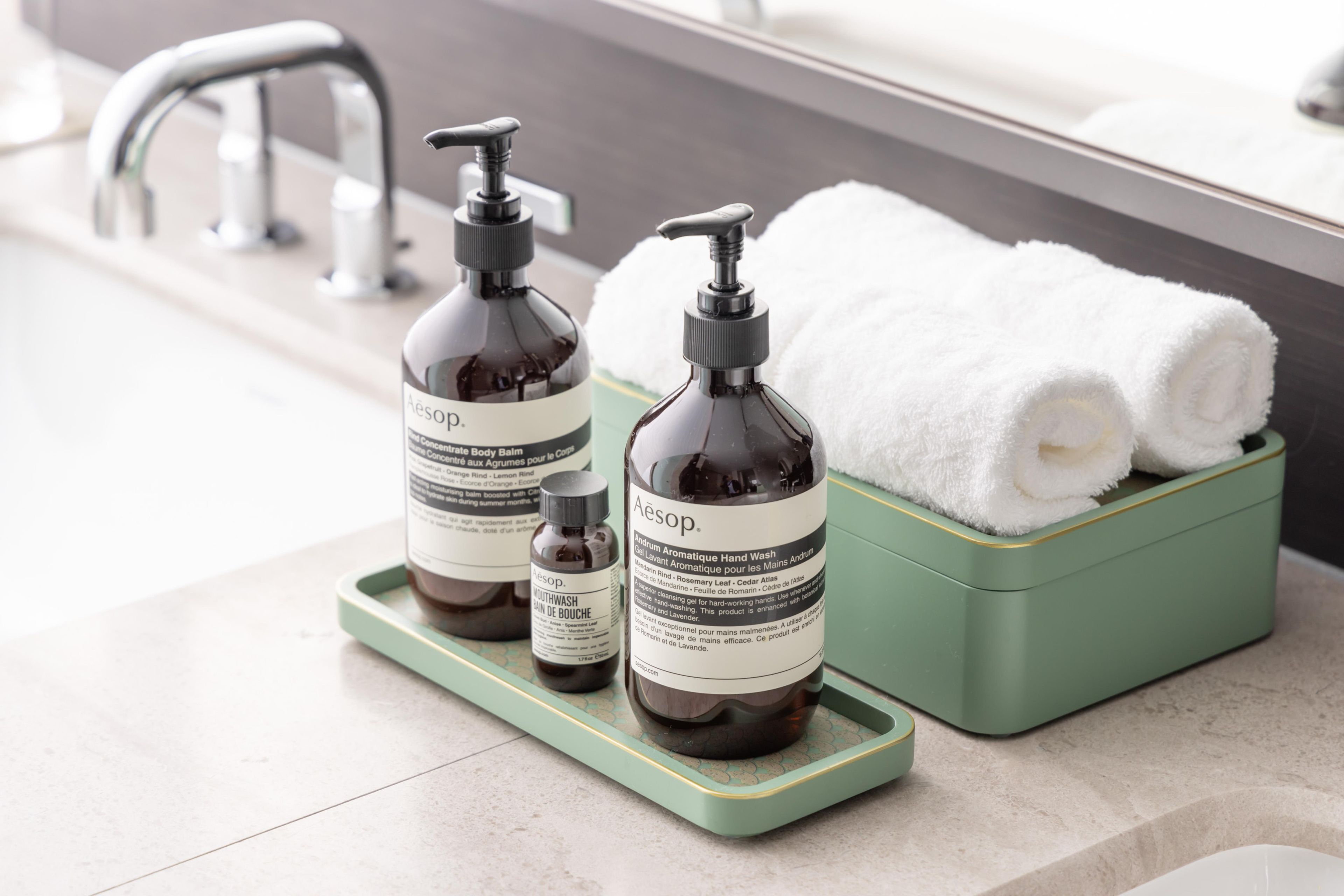 Casa de banho com um saboneteira verde contendo duas embalagens de lavagem de mãos Aesop e uma toalha branca.